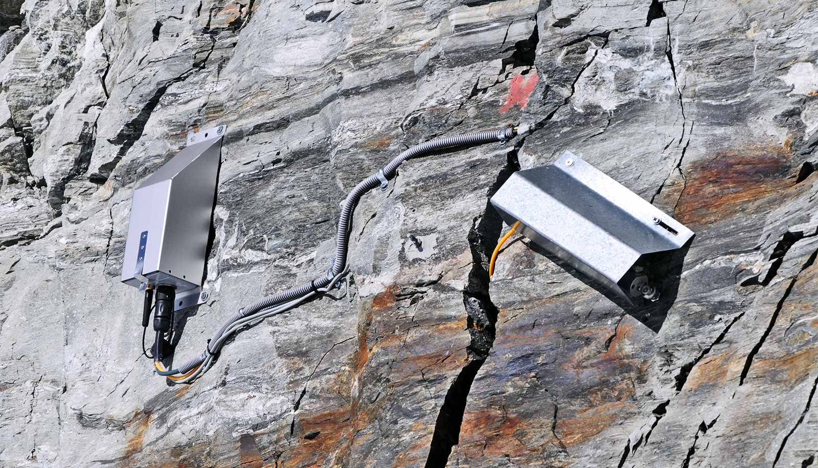 Two metal boxes attached to the rock have wires running into a crack