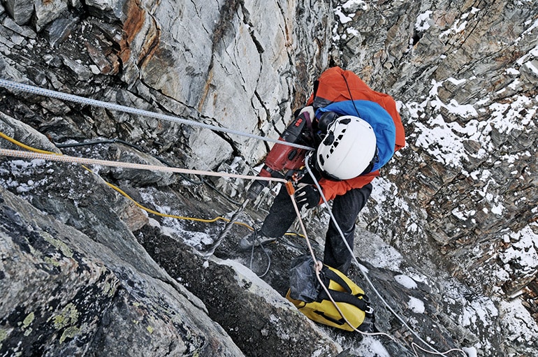 A researcher in climbing gear drills into the rock while hanging from a rope