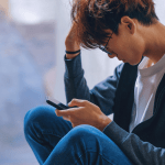 A young man sat down with his head in one hand and holding a phone in the other.