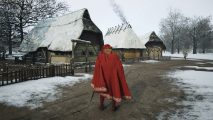 The ruler of the nation, in a red cloak, stands in front of a Manor Lords burgage plot.