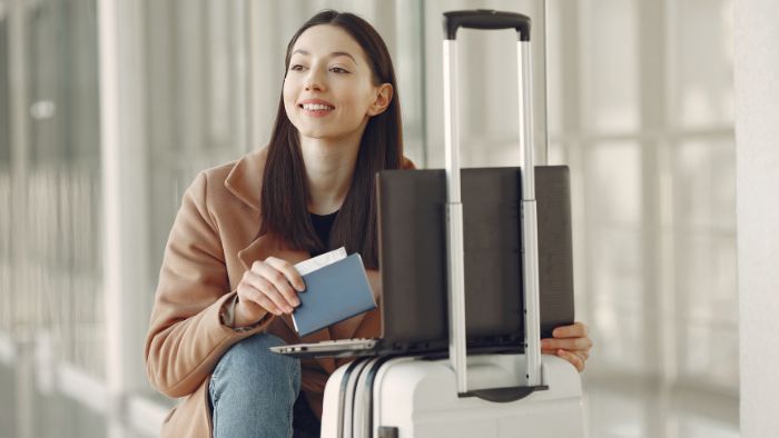 Smiling student holding a passport, bag and suitcase, waiting to travel.