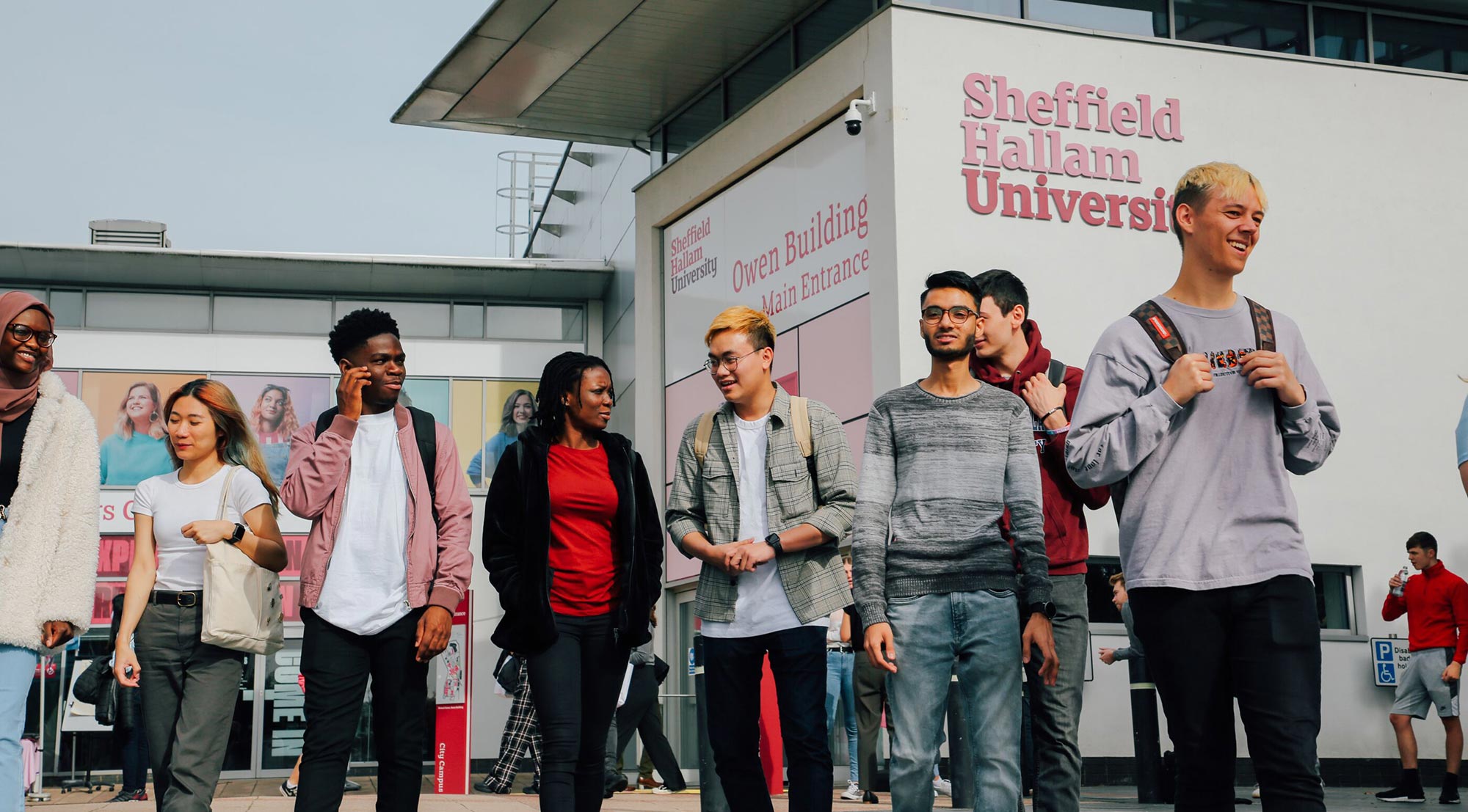 Seven students in front of the Owen Building. They are standing in a line walking forwards, smiling and chatting. 