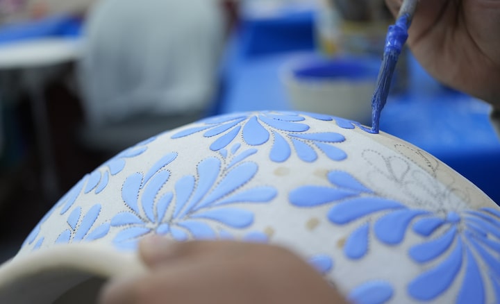 A sample image showing a craftsman adding patterns to a white jar using a brush and blue paint. The focus is on the brush, while the foreground of the jar and the background are blurred