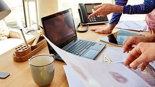 Two colleagues collaborating on a project, with papers and laptops spread out in front of them on a work desk.