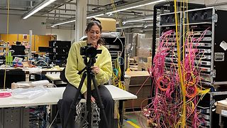 A woman sits behind the camera in a technical room