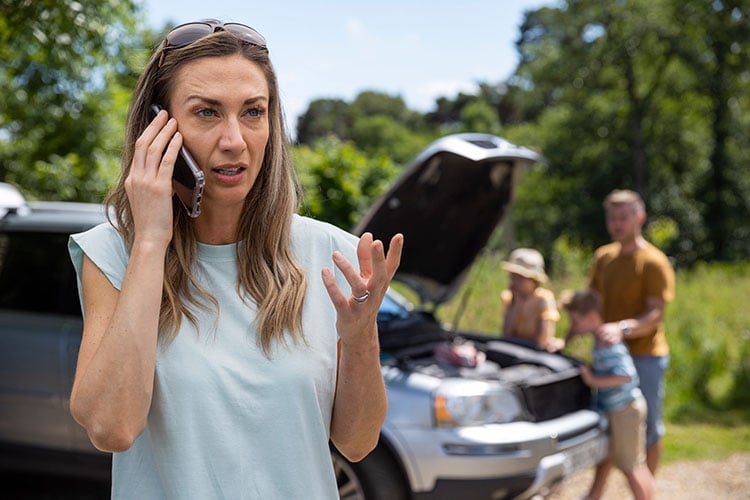 Mid adult woman whose car has broken down on a road trip calls for help with her family seen in the background near the car.