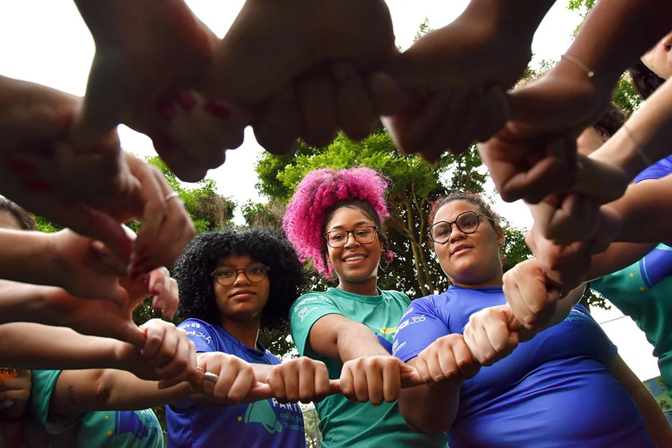 A group of young Brazilian women holding their hands together through their thumbs and with their fists closed.